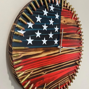 May include: A round wooden clock with an American flag design. The clock face is made of wood with a red, white, and blue paint scheme. The clock hands are silver. The clock is surrounded by a ring of brass bullet casings.