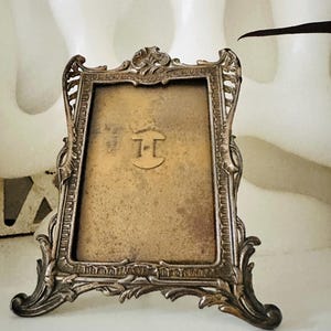 May include: A rectangular metal picture frame with an ornate, dark silver-toned border featuring intricate scrollwork and leaf motifs. The centre contains a weathered, golden-toned backing with a prominent, centred letter H in a serif font.