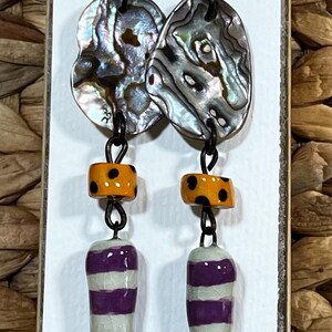 May include: Handmade ceramic earrings with abalone shell accents. The earrings feature orange and black polka dot beads, and white and purple striped ceramic drops. The text on the card reads "SUE SUES ARTFUL JEWELRY HANDMADE CERAMIC ART JEWELRY CREATED BY SUZANNE NOLL."