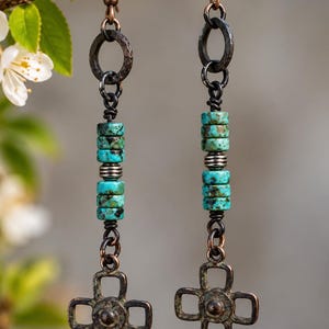 May include: A pair of long dangle earrings featuring dark copper-toned metal hooks and hammered rings. Each earring includes four cylindrical turquoise beads with dark matrix patterns, separated by small silver-toned spacers, ending in a four-lobed metal charm.