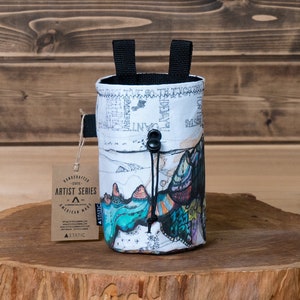 May include: A white chalk bag with a black drawstring closure and two black loops. The bag features a colorful mountain landscape design and a tag that reads "Handcrafted Artist Series American Made". The bag is sitting on a wooden stump.