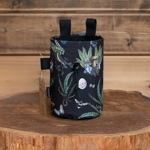 May include: Black fabric bottle holder with a floral and mushroom print. The holder has two black straps and a black drawstring.