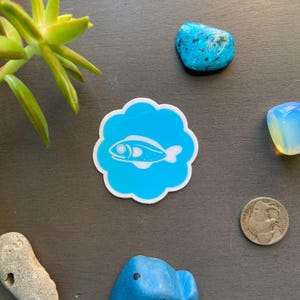 May include: A white fish silhouette sticker on a bright blue background with a white cloud-like border. The sticker is on a gray surface.