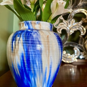 May include: A ceramic vase with a blue, white, and tan drip glaze, holding white flowers with green stems and leaves. The vase is on a dark wooden surface, with a decorative mirror in the background.