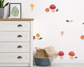 May include: White wall with a variety of red, orange, and yellow mushroom stickers. The mushrooms are different sizes and have white spots. There is a white dresser with four drawers and a wicker basket with a blanket on top of it.