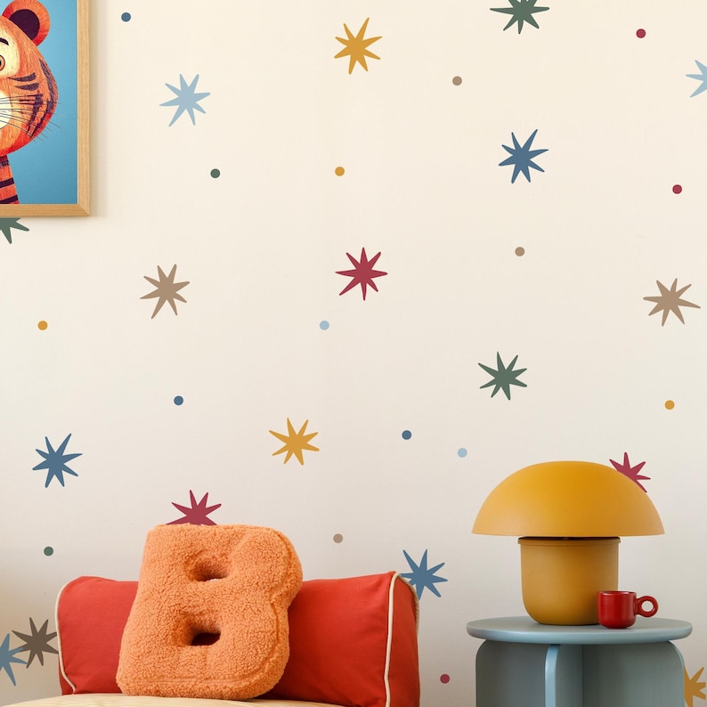 May include: A whimsical children's room scene featuring a wall adorned with colorful star and dot decals. A plush orange letter "B" pillow rests on a red bolster pillow. A mustard-yellow mushroom lamp and a small red mug sit on a blue side table.