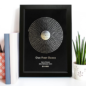 Puede incluir: Impresión enmarcada en negro con un diseño de disco de vinilo plateado. Las ranuras del disco están llenas de letras de canciones. Debajo del disco está el texto "Our First Dance", seguido de nombres y una fecha. Una planta en maceta y libros están en el fondo.