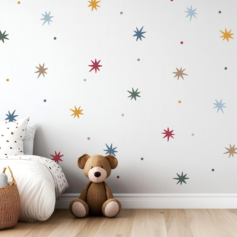 May include: A white wall decorated with colorful star-shaped decals and dots. A brown teddy bear sits on the floor in front of the wall. A bed with a white comforter and a wicker basket are also in the room.