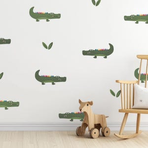 Puede incluir: Una pared blanca decorada con caimanes verdes de dibujos animados con espinas de arcoíris y hojas verdes. Una mecedora de madera y un perro de juguete de madera con ruedas están en primer plano.
