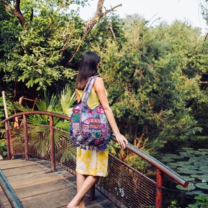 May include: A hobo bag featuring a vibrant patchwork design with intricate embroidery, floral patterns, and blue tassel accents. The bag displays the text Tribe Azure on the front and is carried over the shoulder with a wide, matching fabric strap.