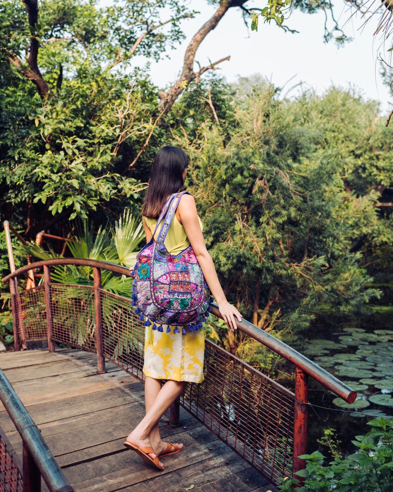 May include: A hobo bag featuring a vibrant patchwork design with intricate embroidery, floral patterns, and blue tassel accents. The bag displays the text Tribe Azure on the front and is carried over the shoulder with a wide, matching fabric strap.