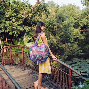 May include: A hobo-style shoulder bag featuring a patchwork design of various floral and geometric fabrics in shades of purple, pink, and blue. The bag is adorned with multiple blue tassels along the bottom edge and displays the text Tribe Azure.