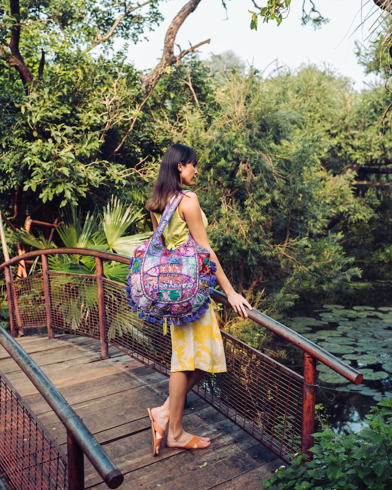 May include: A hobo-style shoulder bag featuring a patchwork design of various floral and geometric fabrics in shades of purple, pink, and blue. The bag is adorned with multiple blue tassels along the bottom edge and displays the text Tribe Azure.
