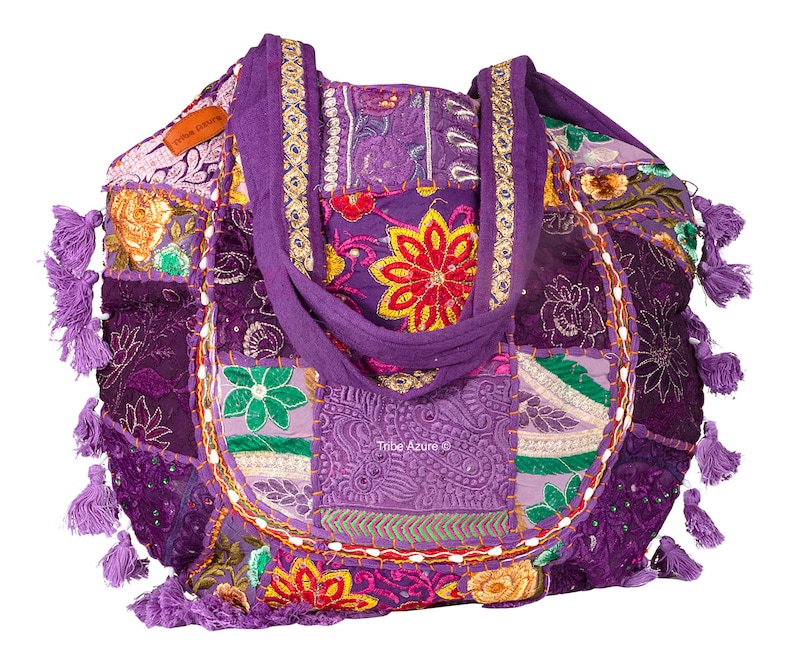 May include: A purple patchwork tote bag featuring embroidered floral patterns, sequins, and decorative stitching. The bag includes two fabric handles, multiple purple tassels along the sides, and a small brown label reading Tribe Azure.