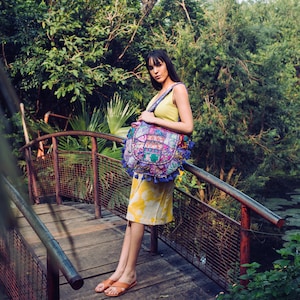 May include: A large, round, multi-colored patchwork shoulder bag featuring intricate embroidery, mirror embellishments, and blue tassel fringe accents. The bag has a wide, patterned fabric shoulder strap and is worn over a yellow sleeveless dress.