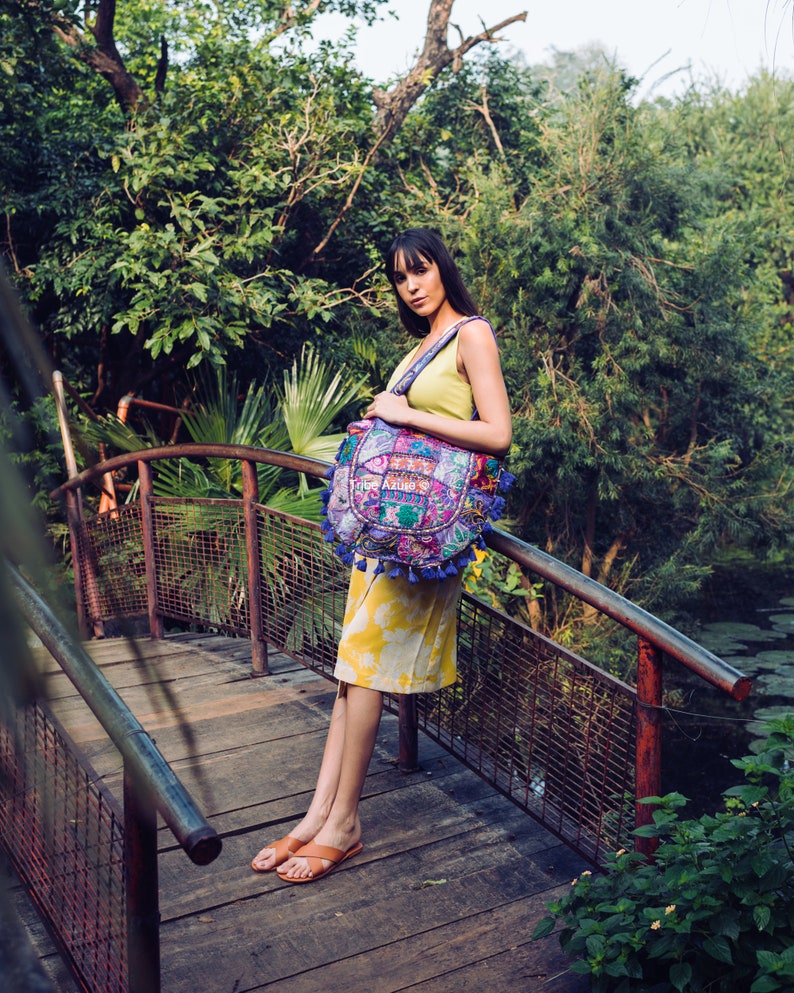 May include: A large, round, multi-colored patchwork shoulder bag featuring intricate embroidery, mirror embellishments, and blue tassel fringe accents. The bag has a wide, patterned fabric shoulder strap and is worn over a yellow sleeveless dress.