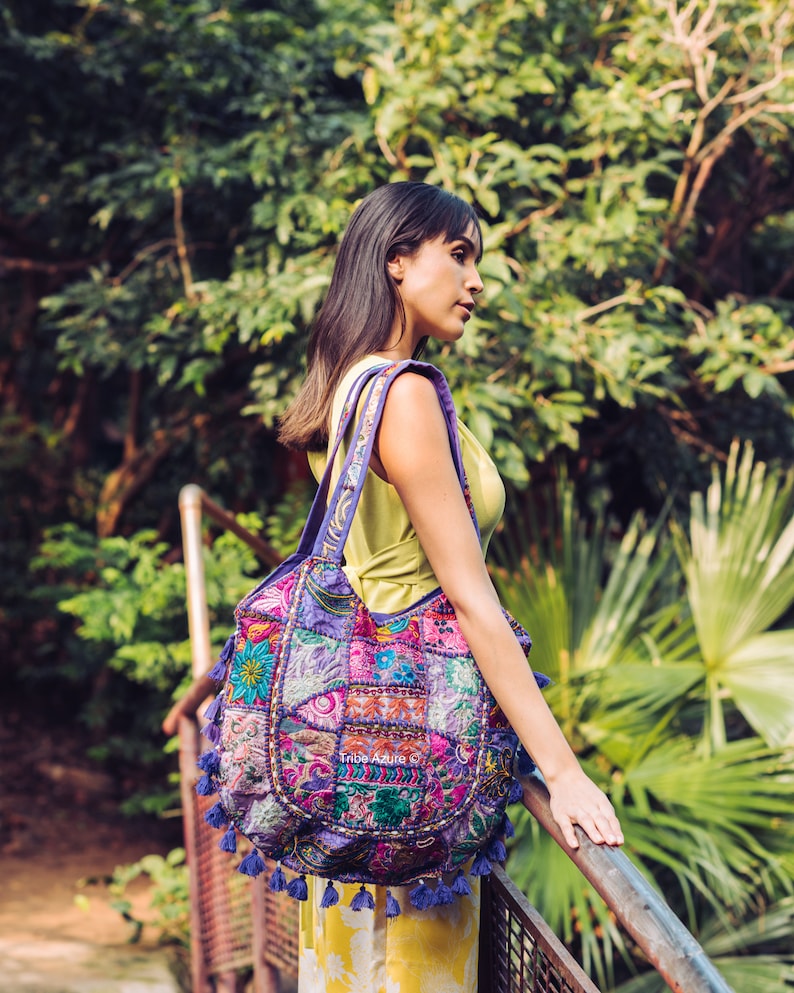 May include: A large hobo-style shoulder bag featuring a vibrant patchwork design with intricate embroidery in shades of purple, pink, blue, and green. The bag is adorned with numerous small purple tassels along the bottom and sides, with the text Tribe Azure.