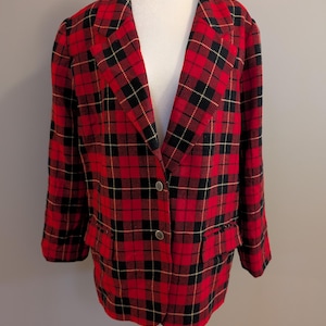 Op de afbeelding: Een rood, zwart en goud geruit jasje met twee voorzakken en twee zilveren knopen. Het jasje wordt gepresenteerd op een witte mannequin. Het ruitpatroon is een klassiek ontwerp.