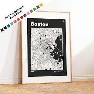 May include: A black and white map print of Boston, Massachusetts, with the city name in white text at the top. The map shows the city's streets, waterways, and surrounding areas. The print is framed in a light wood frame and is hanging on a white wall.