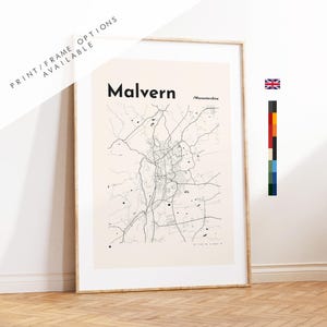 May include: A framed wall art print featuring a minimalist black line map of Malvern, Worcestershire on a light beige background. The text Malvern and Worcestershire are printed in black. A small Union Jack flag and a vertical color palette are visible.