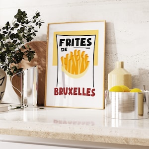 May include: A vintage-style poster with a yellow background and black text that reads "Frites de Bruxelles" with an illustration of french fries. The poster is framed in a gold frame and is hanging on a white wall.