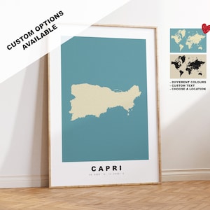 Puede incluir: Una impresión enmarcada de un mapa de Capri, Italia, con el contorno de la isla en beige sobre un fondo azul verdoso. La impresión incluye el nombre "Capri" y las coordenadas "40.5533° N, 14.2222° E". La impresión está enmarcada en un marco de madera claro. La imagen también incluye un mapa del mundo con un alfiler de corazón rojo, y un mapa del mundo en blanco y negro.