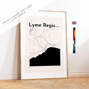May include: A framed wall art print featuring a minimalist map of Lyme Regis, Dorset. The design displays black line work for roads and a solid black shape for the coastline against a light beige background. The text Lyme Regis and Dorset is visible.