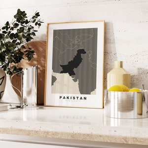 May include: A framed print of a map of Pakistan with the country name printed below the map. The map is in black and white with a gray background. The frame is a light wood color. The print is sitting on a white countertop with a silver pitcher and a silver bowl with lemons.
