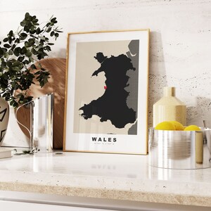 May include: A framed print of a map of Wales with the text "WALES" below the map. The map is black and white with a red dot in the center. The frame is gold and the background is white.