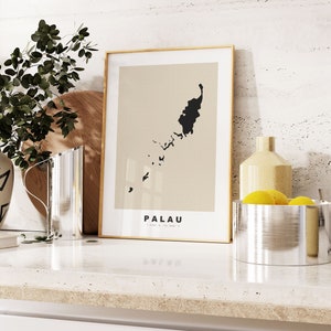 May include: A framed print of a map of Palau with the word "PALAU" printed below the map. The print is on a white background and is framed in a light wood frame. The print is sitting on a white countertop with a vase of greenery and a silver bowl with lemons in the background.
