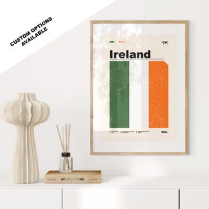 Puede incluir: Un póster enmarcado con un diseño minimalista que presenta la bandera de Irlanda. El póster tiene un fondo blanco con un diseño de rayas verticales verdes y naranjas. La palabra "Irlanda" está impresa en negro en la parte superior del póster. El póster está enmarcado en un marco de madera claro.