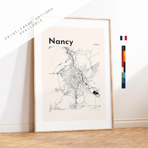 May include: Framed print of a map of Nancy, France, in black and white. The city name is displayed in a bold font at the top, with the country name below. The print is in a light-colored frame and leans against a white wall.