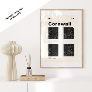 May include: A framed print with a black and white graphic design of the flag of Cornwall. The print has a beige background with the word "Cornwall" in black text at the top. The print is framed in a light wood frame.