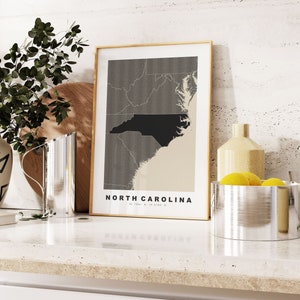 May include: A framed print of a map of North Carolina with the state name in a black and white font. The map is on a beige background with a black outline of the state. The print is sitting on a white countertop with a silver pitcher and a silver bowl with lemons.