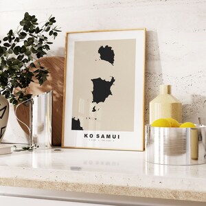 Könnte beinhalten: Ein gerahmter Druck mit beigefarbenem Hintergrund und einer schwarzen Silhouette der Insel Koh Samui, Thailand. Der Druck trägt den Titel "Koh Samui" und wird auf einer weißen Arbeitsplatte mit einer silbernen Kanne und einer silbernen Schüssel gefüllt mit gelben Zitronen gezeigt.