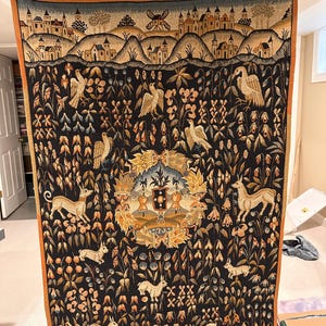 May include: A rectangular woven tapestry featuring a dark background with a central heraldic emblem surrounded by various animals including deer, rabbits, and birds amidst floral motifs. The top border depicts a landscape with buildings and a windmill.