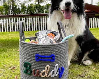 Op de afbeelding: Een grijze touwmand met de naam "Buddy" in kleurrijke letters en een blauwe botvormige applicatie. De mand is gevuld met hondenspeelgoed en -snoepjes. Een zwart-witte Border Collie zit naast de mand op een grasveld.