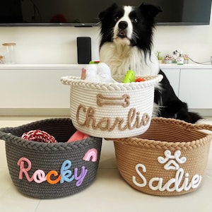 May include: Three woven rope baskets in white, gray, and tan, each with a pet's name embroidered on the front. The white basket has "Charlie" embroidered, the gray basket has "Rocky", and the tan basket has "Sadie". A black and white dog is in the background.