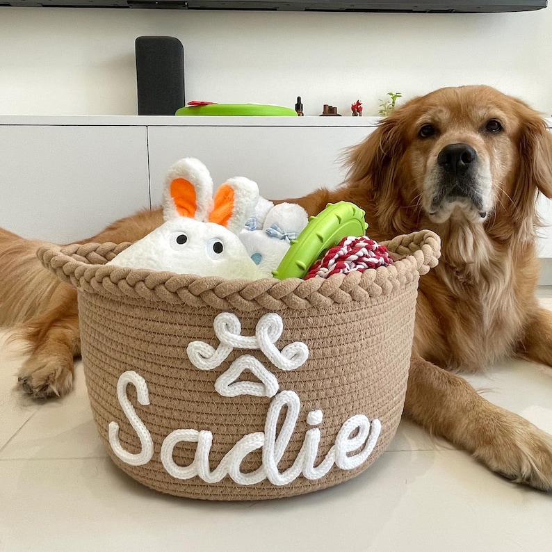May include: A woven brown pet toy basket with the name "Sadie" in white script. The basket is filled with plush toys, including a white bunny with orange ears, a blue toy, and a green chew toy. A golden retriever dog is in the background.