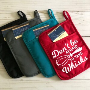 May include: Four pot holders in different colors. The red pot holder has the text "Don't be afraid to take whisks" with a whisk graphic. The other pot holders are gray, black, and teal.