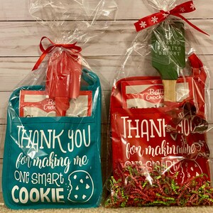 May include: Two gift bags filled with baking supplies. The blue bag has a cookie cutter and a bag of baking mix. The red bag has a spatula, a cookie cutter, and red and green shredded paper. The blue bag has the text "Thank you for making me one smart cookie". The red bag has the text "Thank you for making me one smart cookie".