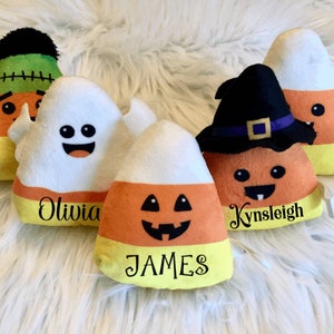 May include: Five Halloween-themed candy corn plush toys with embroidered faces and names. The toys are:  Olivia, James, Kynsleigh, a ghost, and a Frankenstein.