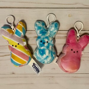 May include: Three plush Peep bunny keychains. One is pink with a tie-dye pattern, one is blue with a tie-dye pattern, and one is yellow, pink, and blue with a striped pattern. The keychains have the word "Peeps" on the tag.