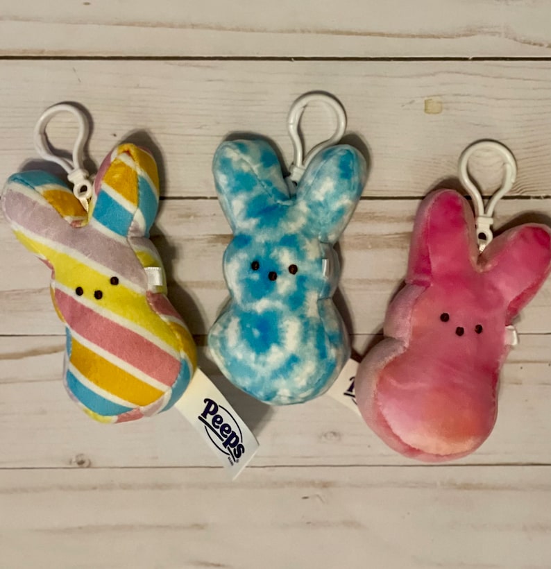 May include: Three plush Peep bunny keychains. One is pink with a tie-dye pattern, one is blue with a tie-dye pattern, and one is yellow, pink, and blue with a striped pattern. The keychains have the word "Peeps" on the tag.