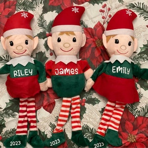 May include: Three personalized Christmas elf dolls with red and green outfits. The elves are named Riley, James, and Emily. Each elf has a red and white striped stocking and green shoes.