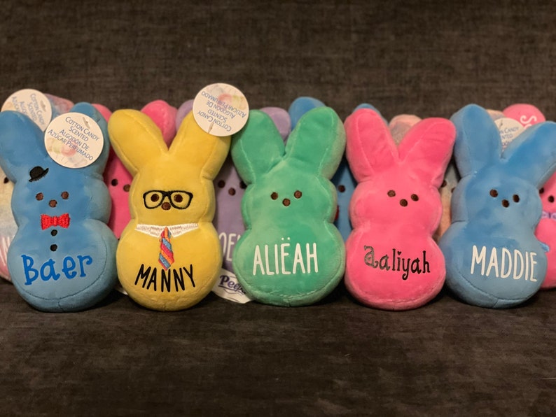 May include: A row of seven plush Easter bunny toys, each with a different color and personalized name. The names are Baer, Manny, Alie&aacute;h, @aliyah, Maddie, and two others. The bunnies are all wearing different accessories, such as glasses, a tie, and a bow tie.