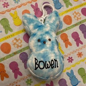 May include: A blue and white tie-dye plush bunny with a black embroidered name "Bowen" on the front. The bunny is attached to a white plastic key chain.