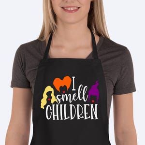 May include: Black apron with the text "I smell CHILDREN" in white script. The apron features cartoon illustrations of three witches' hair in yellow, orange, and purple. The apron has a neck loop and waist ties.