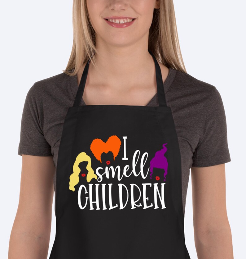 May include: Black apron with the text "I smell CHILDREN" in white script. The apron features cartoon illustrations of three witches' hair in yellow, orange, and purple. The apron has a neck loop and waist ties.