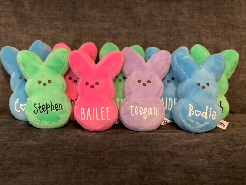 May include: Seven plush Peeps bunny shaped Easter decorations with personalized names. The Peeps are in the colors green, pink, purple, and blue.